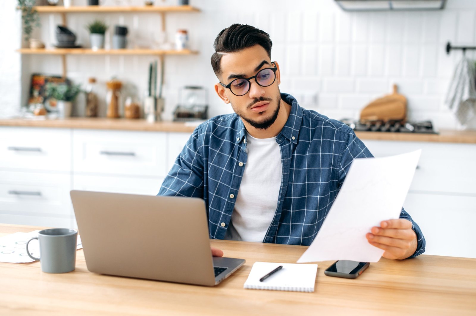 Focused serious arabian or indian guy with glasses, freelancer or office worker working remotely, sit at a desk at home in kitchen, working on a project uses laptop, concentrated analyzing documents